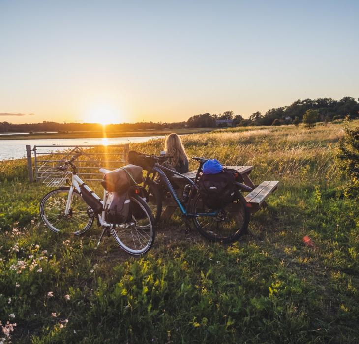 Kvinne slapper av ved et bord og en benk, midt på et jorde, og beundrer soloppgangen over vannet ved knudshoved på Fyn